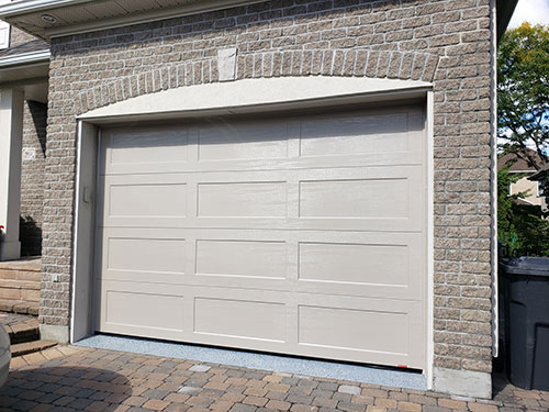 Une porte de garage beige à panneaux horizontaux, installée dans un mur de briques en pierre, à côté d'une allée pavée.