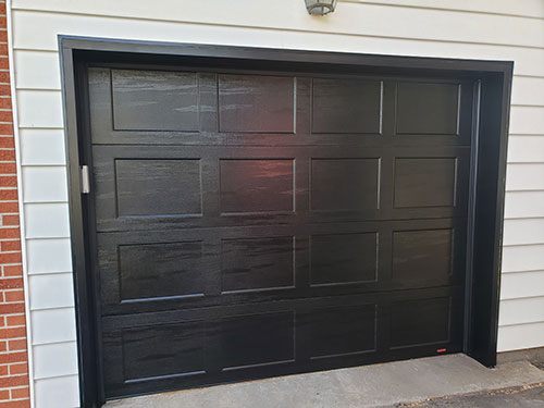 Porte de garage noire moderne avec un design à panneaux, encadrée par un mur blanc et un parement en brique.