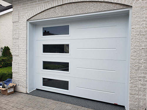 Porte de garage gris clair moderne avec quatre fenêtres horizontales, sur un mur de pierre texturée et une allée pavée.