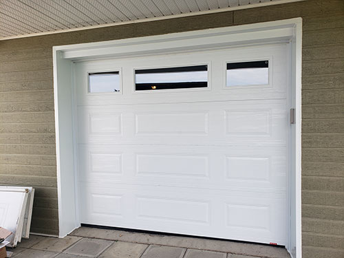 Porte de garage blanche avec trois fenêtres rectangulaires en haut, posée sur un mur gris texturé entouré de pavés.