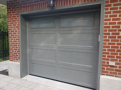 Porte de garage grise moderne avec panneaux horizontaux et fenêtres, encadrée par un mur de briques et une allée pavée.