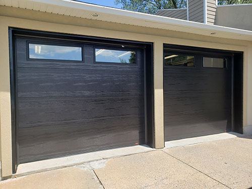 Deux portes de garage modernes de couleur noire avec une texture de bois horizontale et de petites fenêtres, placées contre un mur de bâtiment de couleur claire.