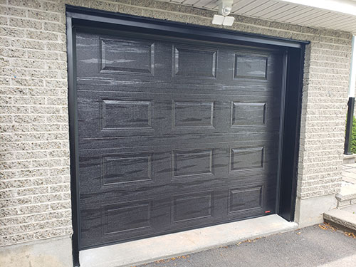 Gros plan sur une porte de garage noire moderne avec un design à panneaux texturés, contre un mur en briques gris clair.