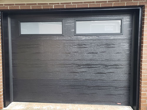Porte de garage noire avec deux fenêtres rectangulaires en haut, panneaux texturés horizontaux sur fond de mur en briques.