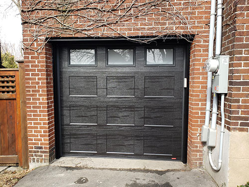 Porte de garage noire moderne avec un design texturé, encadrée par des murs en briques rouges, des tuyaux et une clôture en bois.
