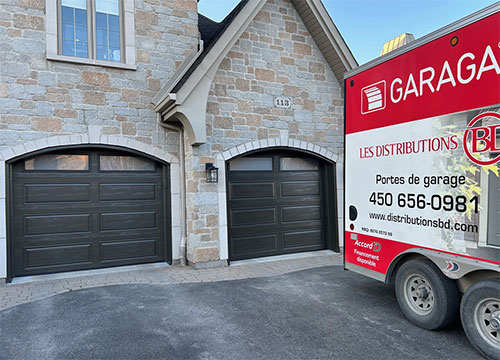 Deux portes de garage sombres sur une maison en façade de pierre, avec un camion de livraison stationné à proximité.