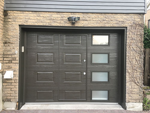 Porte de garage brun foncé moderne avec trois fenêtres rectangulaires, panneaux texturés, luminaire au-dessus, encadrée par des briques.