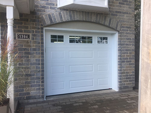 Porte de garage blanche avec fenêtres décoratives, installée dans une arche en briques, à côté d’un sentier en pierre.