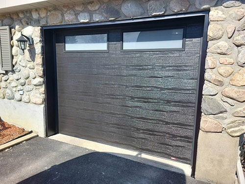 Porte de garage noire avec deux fenêtres étroites, encadrée par un mur en pierre, dans une allée résidentielle.