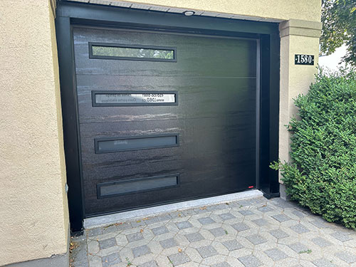 Porte de garage noire moderne avec trois fenêtres horizontales, sur une façade de maison claire entourée de verdure.