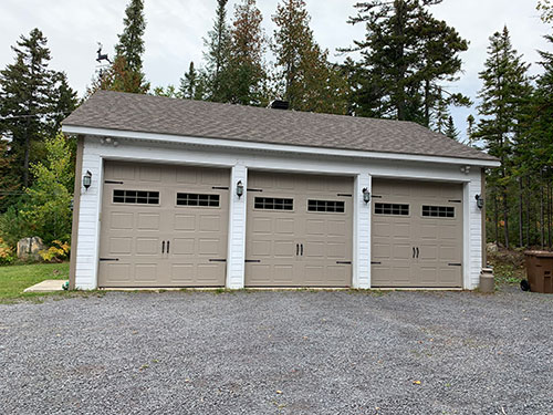 Garage triple avec portes beiges et lanternes, dans une allée en gravier entourée d'arbres verts dans un cadre paisible.
