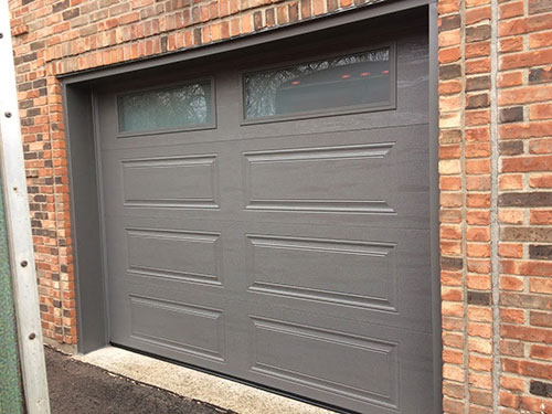 Porte de garage grise moderne avec deux petites fenêtres, contre un mur en briques texturé au design contemporain.