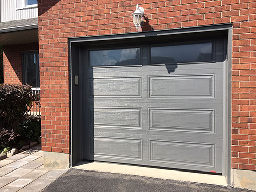 Porte de garage grise avec panneaux décoratifs, dans un mur en briques, avec un luminaire extérieur et un chemin bien entretenu.
