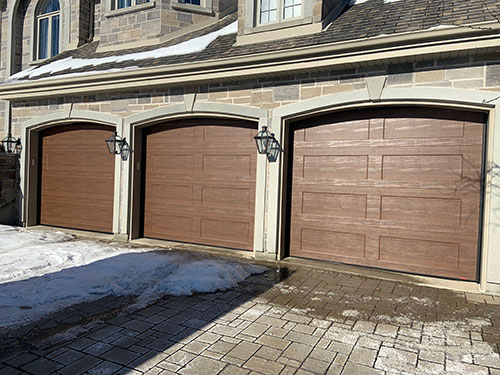 Trois portes de garage en bois moderne avec panneaux horizontaux, encadrées par des murs en pierre et lanternes vintage, sur une allée enneigée.