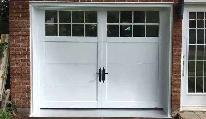 Porte de garage blanche avec fenêtres décoratives, encadrée par un mur en briques, à côté d’une porte vitrée.