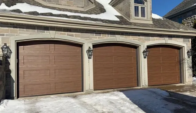 Trois portes de garage en bois moderne avec panneaux horizontaux, encadrées par des murs en pierre et lanternes vintage, sur une allée enneigée.