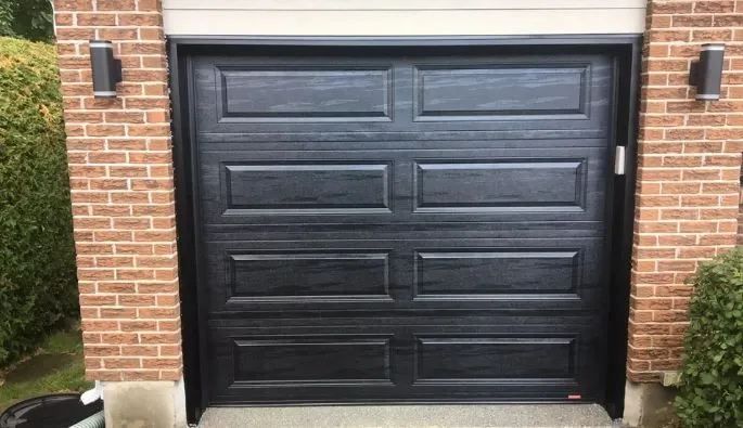 Gros plan sur une porte de garage noire moderne avec un design à panneaux texturés, contre un mur en briques.